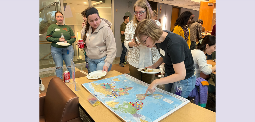 Three people stand around a cork board map displayed on a table. They are examining which places have been pinned. 