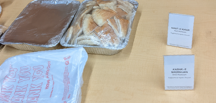 A aluminum food container sits on a wooden table next to another aluminum food container in a "thank you plastic bag" and a container of fresh pita bread 