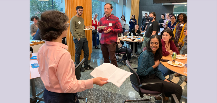Indira Raman stands in the center of a room holding a paper. She addresses the room and people seated at round tables and standing in the space listen on. 
